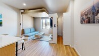 Located at 319 N Pottstown Pike in Exton, PA, this efficiently designed single-floor unit maximizes every square foot through smart layout planning and modern design choices. Light hardwood flooring runs throughout, creating a warm, cohesive aesthetic from the entrance corridor to the living area.

The entrance hallway leads into the heart of the unit: an open-plan kitchen and living room. The kitchen is outfitted with flat-front light wood cabinetry, stainless steel appliances including a built-in microwave, range, and refrigerator, and a crisp white quartz peninsula with an integrated sink and bar seating—ideal for dining or working. The living area provides comfortable space for a sofa and lounge furniture, with a window allowing natural light to flow in.

A standout feature is the elevated loft platform above the living space, which accommodates a sleeping area and makes inventive use of vertical space. Built-in shelving and a closet provide practical storage solutions throughout.

The bathroom is clean and minimal, featuring light wood-toned cabinetry, light hardwood flooring, and a shower area alongside a full toilet suite. An in-unit laundry area adds further convenience. The overall palette—white walls, light wood tones, and stainless accents—gives the space a bright, contemporary feel.