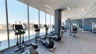 This well-appointed fitness center offers an impressive workout environment defined by its expansive floor-to-ceiling glass walls, which flood the space with abundant natural light and frame sweeping panoramic views of the surrounding cityscape. The open layout accommodates a generous selection of Life Fitness cardio equipment, including elliptical trainers and upright bikes, all positioned to take full advantage of the outdoor views. Along the opposite wall, a bank of mirrors visually expands the space while reflecting the bright, airy atmosphere. Strength-training and cable machine equipment is arranged along the perimeter, providing a well-rounded fitness experience. The flooring consists of durable, slate-toned rubber tiles suited for high-traffic gym use. Recessed linear lighting complements the natural light, ensuring consistent illumination throughout. Structural columns with a clean, gray finish contribute to the contemporary aesthetic. The overall design is sleek and minimalist, combining functionality with a polished, modern ambiance that makes the most of its elevated position within the building.