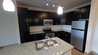 The kitchen is a standout feature of this single-floor space, showcasing a well-designed layout that balances functionality and modern style. Dark espresso-toned cabinetry lines the walls from base to upper levels, providing generous storage while contributing to the space's bold, contemporary aesthetic. The cabinetry is complemented by light-toned granite countertops with subtle veining, creating a striking contrast that adds visual interest throughout the kitchen.

A central island serves as the focal point, equipped with a double-basin stainless steel sink and matching granite surface, offering additional workspace and a natural gathering point. Stainless steel appliances, including a range with oven, over-the-range microwave, and refrigerator, maintain a cohesive, professional look.

Lighting is thoughtfully layered, combining a track lighting system along the ceiling with a sleek pendant light suspended above the island, ensuring the workspace is well-lit. A white subway tile backsplash provides a clean, classic backdrop between the countertops and upper cabinets. Light hardwood flooring grounds the space, adding warmth that balances the darker tones of the cabinetry.