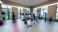 This fitness center offers a comprehensive workout environment thoughtfully designed with both functionality and comfort in mind. The space is bathed in natural light, courtesy of expansive floor-to-ceiling windows that line two walls, providing open views of the surrounding outdoor area, including landscaped grounds and adjacent buildings. The bright, airy atmosphere is further enhanced by recessed lighting along the ceiling perimeter, ensuring consistent illumination throughout.

The layout accommodates a well-rounded selection of exercise equipment. Cardio machines, including multiple treadmills and an elliptical trainer, are positioned to take advantage of the natural light and outdoor views. Strength training equipment, including a weight bench and cable machine, occupies the remaining floor space, offering users a variety of resistance workout options. A flat-screen television is mounted on the wall, providing entertainment during workouts.

The flooring transitions between a neutral tone and red rubberized matting beneath the cardio equipment, offering both durability and support. Wall-mounted sconces add a subtle decorative touch, contributing to the clean, modern aesthetic of the space.