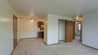 This single-floor space features an open-concept layout that seamlessly connects the living area to the kitchen. The living space is finished with wall-to-wall carpet in a neutral tone, creating a warm and comfortable atmosphere throughout. A ceiling fan with an integrated light fixture is centrally positioned, providing both airflow and ambient lighting to the main living area.

A partial dividing wall separates the living space from a built-in closet, which is fitted with a hanging rod and open shelving, offering practical storage. An additional door is visible beyond the closet, suggesting access to a separate room or hallway.

The kitchen is tucked into the background and features dark wood cabinetry, a countertop workspace, and a black range, providing functional cooking essentials in a compact footprint. The cabinetry extends to upper wall-mounted units, maximizing vertical storage.

Throughout the space, neutral beige walls complement the warm wood tones of the doors and cabinetry. A textured ceiling adds subtle detail overhead. The overall ambiance is practical and understated, with a cohesive, neutral palette that offers a versatile backdrop for furnishing and personalization.