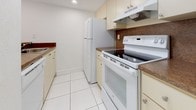 This single-floor residence is designed with a clean, neutral aesthetic throughout, characterized by white walls and an abundance of natural light. The open living area features large-format white ceramic tile flooring and is anchored by a pendant light fixture. A half-wall breakfast bar separates the living space from the galley-style kitchen, which is equipped with cream cabinetry, dark laminate countertops, a white refrigerator, an electric range, and a range hood. Recessed lighting illuminates the kitchen area. A sliding glass door in the living room provides direct access to an outdoor area and frames views of surrounding greenery, enhancing the sense of openness. Louvered bifold closet doors are found in multiple areas, offering practical storage solutions. The bedrooms are carpeted in a neutral beige tone and each features vertical blind-covered windows that allow for natural light while maintaining privacy. The bathroom includes a tiled tub-shower combination enclosed by sliding glass doors, accented with a decorative mosaic tile border. The hallway and entry areas continue the white tile flooring, maintaining a cohesive look throughout the unit.