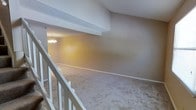 This two-story residential space offers a straightforward and functional layout spread across two levels connected by a staircase with clean white railings. The upper level provides an open view down to the main living area below, creating a sense of vertical spaciousness throughout. The lower level features wall-to-wall carpeting in a neutral tone that contributes to a warm and comfortable atmosphere. A classic chandelier serves as a focal point in the lower living area, adding a touch of elegance to the otherwise understated interior. The walls are finished in a muted, earthy tone that complements the light carpeting and white trim throughout. Textured ceilings add a subtle architectural detail to each level. The open sightlines from the upper landing to the lower living space enhance the overall sense of openness, making the home feel connected and cohesive. The neutral color palette and carpeted floors create a welcoming environment suited to a variety of furnishing styles. The layout across two floors provides natural separation between living and private spaces, offering both functionality and comfort for everyday living.