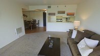 This single-floor residence is designed with an open-concept layout that seamlessly connects the main living areas. The living room serves as the central gathering space, featuring wall-to-wall carpeting that adds warmth underfoot. A dark wood coffee table anchors the seating area, which includes a cushioned sofa positioned to take advantage of the open sightlines throughout the space.

A pass-through counter connects the living area directly to the kitchen, enhancing the open feel while maintaining a defined cooking zone. The kitchen is equipped with a range, sink, and overhead shelving — the latter decorated with playful café-themed signage that adds a touch of personality to the space. Light-toned cabinetry and countertops keep the kitchen feeling bright and clean.

Adjacent to the living room, the dining area accommodates a full table and chair set on hardwood-style flooring, providing a visual contrast to the carpeted living space. The transition between flooring materials subtly delineates each functional zone. Overall, the layout prioritizes practicality and flow, making efficient use of the available space across the single floor.