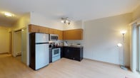 This single-floor unit showcases a well-designed open-concept kitchen that serves as the focal point of the space. The kitchen features a two-toned cabinetry design, combining warm honey-toned upper cabinets with sleek dark lower cabinets, creating a modern and visually appealing contrast. Full-size appliances, including a refrigerator, electric range, and built-in microwave, are thoughtfully integrated into the layout, maximizing both functionality and space efficiency. The L-shaped counter configuration provides ample workspace and includes a sink station along the far wall. Track lighting mounted on the ceiling provides targeted illumination throughout the kitchen area. Light hardwood-style flooring extends throughout the open living area, lending warmth and continuity to the space. The neutral white walls and clean lines contribute to a bright, airy atmosphere. The open layout between the kitchen and living area creates a sense of spaciousness, making the unit feel larger than its footprint. Overall, the space balances practical design with contemporary aesthetics, offering a functional and comfortable living environment.