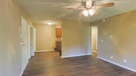 This single-floor space offers an open-concept layout that seamlessly connects the living area to the kitchen. Dark hardwood flooring runs throughout, providing a warm, cohesive foundation that contrasts cleanly with the neutral beige-toned walls and bright white baseboards. The living area is centered beneath a multi-blade ceiling fan with integrated lighting, ensuring both comfort and illumination. The kitchen is partially open to the main living space and features warm wood cabinetry, a dark countertop, and a stainless steel appliance, with upper and lower cabinet storage offering practical functionality. A doorway visible to the right suggests access to additional rooms, maintaining a natural flow through the unit. The layout benefits from multiple light sources, including flush-mount ceiling fixtures. The overall ambiance is understated and functional, with clean lines and a neutral palette that creates a versatile backdrop suitable for a variety of interior styles. The open sightlines between the living and kitchen areas enhance the sense of spaciousness throughout the single-level floor plan.