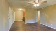 This single-floor space offers an open-concept layout that seamlessly connects the living area to the kitchen. Dark hardwood flooring runs throughout, providing a warm, cohesive foundation that contrasts cleanly with the neutral beige-toned walls and bright white baseboards. The living area is centered beneath a multi-blade ceiling fan with integrated lighting, ensuring both comfort and illumination. The kitchen is partially open to the main living space and features warm wood cabinetry, a dark countertop, and a stainless steel appliance, with upper and lower cabinet storage offering practical functionality. A doorway visible to the right suggests access to additional rooms, maintaining a natural flow through the unit. The layout benefits from multiple light sources, including flush-mount ceiling fixtures. The overall ambiance is understated and functional, with clean lines and a neutral palette that creates a versatile backdrop suitable for a variety of interior styles. The open sightlines between the living and kitchen areas enhance the sense of spaciousness throughout the single-level floor plan.