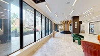 Cosmo440's ground-floor lobby and amenity level in Newark, NJ, is a thoughtfully designed space that blends contemporary architecture with warm, inviting details. The expansive floor plan is anchored by a striking sculptural centerpiece — a dramatic golden wood-slat column rising from a circular bench seat — creating an immediate focal point upon entry. Large floor-to-ceiling windows line the exterior walls, flooding the polished marble-tile floors with abundant natural light and offering clear views to the surrounding streetscape.

The lobby transitions seamlessly into multiple distinct zones. A stylish lounge area features bold, sculptural velvet seating in rich jewel tones — deep blue, burnt orange, and forest green — arranged around a patterned ottoman, adding a vibrant, curated aesthetic. Nearby, an elevator bank is framed by a dramatic geometric LED light installation set against dark accent walls.

A co-working and café-style area offers polished concrete ceilings with exposed ductwork and Edison bulb pendants, lending an industrial contrast to the otherwise refined setting. Leather and wood furnishings, bar-height seating, and collaborative tables make it a functional workspace. A separate lounge zone features darker tones, hanging greenery, and a wet bar with counter seating. Clean, well-lit corridors connect to glass-enclosed meeting rooms, completing this versatile and design-forward amenity floor.