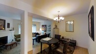 This two-floor space is thoughtfully designed to support a productive professional environment. The main office area is anchored by a dark, circular meeting table surrounded by leather upholstered chairs, ideal for consultations or collaborative sessions. A decorative multi-arm chandelier with frosted glass shades serves as a refined focal point, casting warm, inviting light throughout the room. A framed mirror on the wall adds depth and brightness to the space.

An arched passageway connects the front area to an inner office, where a dedicated workstation is set up with a desktop computer, a desk lamp, and organized work surfaces along the walls. Framed artwork and a collection of small frames add a personalized touch, while a tall filing cabinet provides practical storage. Natural light filters in through a window, enhancing the welcoming atmosphere of the inner room.

The flooring transitions between warm hardwood and carpeted areas, defining the different functional zones. The walls are finished in soft, neutral tones, contributing to a calm and focused ambiance throughout. The overall layout of the space balances professionalism with comfort, making it well-suited for office or business use across both floors.