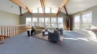 This single-floor space makes a striking architectural impression with its dramatic vaulted ceilings, where exposed natural wood beams intersect against crisp white surfaces, creating a bold structural statement overhead. The open-plan layout is generously proportioned, offering a sense of expansiveness that is further enhanced by the soaring roofline and the volume of space it creates.

A wall of large, multi-pane windows lines one side of the room, flooding the interior with natural light and offering unobstructed views of the surrounding outdoor environment. The soft grey carpeting throughout lends a comfortable, cohesive feel to the space, complemented by sage-green painted walls that add warmth and a calming ambiance.

The lounge area is thoughtfully arranged with a mix of upholstered seating — including a sofa and coordinating armchairs — centered around a round wooden coffee table atop a neutral area rug. A wooden bookshelf and side tables complete the furnishings. A railed balcony-style staircase opening adds architectural interest, and a small counter or service bar area is visible toward the rear of the space. Emergency exit doors provide access at the front, suggesting a community or shared-use facility. Overall, the space balances practicality with an inviting, airy aesthetic.