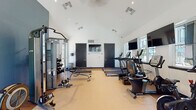 This single-level fitness center is designed to support a comprehensive workout experience within a bright, well-appointed space. The room features a striking vaulted ceiling that rises to a considerable height, lending the interior an open, airy quality that enhances the overall sense of spaciousness. White walls contrast with dark-framed doors and a bold blue accent wall, giving the space a clean, modern aesthetic.

The facility is equipped with a full lineup of TRUE-brand cardio equipment, including treadmills, elliptical trainers, and a stationary bike, all positioned along the windowed wall to allow natural light and outdoor views during workouts. Multiple flat-screen televisions are mounted above the cardio stations for entertainment. On the strength side, the space includes a multi-function cable machine, adjustable weight benches, and a tiered dumbbell rack with a wide range of weights, accompanied by a large wall mirror that enhances visibility and the sense of depth.

Recessed lighting supplements the natural light provided by multiple windows along the perimeter. A set of glass double doors at one end opens directly to the outdoor courtyard, further connecting the interior to the surrounding environment. The flooring is a durable, light-toned fitness surface suited for both cardio and strength training activities.