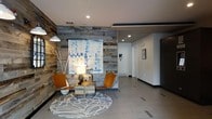 This single-floor space makes a strong first impression with its thoughtfully designed lobby area. The standout feature is a striking reclaimed wood accent wall, composed of weathered planks in varying shades of grey and brown, lending the space a warm yet contemporary rustic character. Against this textured backdrop, a vibrant blue-and-white abstract artwork serves as a bold focal point, adding a splash of color and artistic flair to the environment.

The seating area is curated with a pair of mid-century modern-style amber leather chairs arranged around a low wooden table, all anchored by a circular patterned area rug. An industrial-style pendant lamp adds to the eclectic, design-forward aesthetic of the space.

The flooring throughout consists of large-format light grey tiles, contributing to a clean, open feel. The white ceiling is fitted with recessed lighting that provides even, ambient illumination across the space. A corridor extending from the main area hints at additional rooms or offices beyond, framed by clean white walls and accented with framed artwork. Overall, the space balances industrial and modern design elements to create an inviting and professional atmosphere.