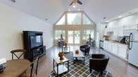 This single-story space is defined by its dramatic vaulted ceiling, which rises to a striking peak and is complemented by a large triangular window arrangement at the front wall. The expansive glazing floods the interior with natural light, creating a bright and welcoming atmosphere throughout. Dark hardwood flooring runs across the main area, providing a warm contrast to the crisp white walls and trim.

The main lounge area is furnished with dark leather accent chairs arranged around low wooden coffee and side tables, all set atop a patterned area rug that anchors the seating zone. A large flat-screen television on a dark media console anchors one side of the room. A ceiling fan with a light fixture is centrally positioned to circulate air throughout the open volume.

Toward the rear, glass double doors lead to an outdoor area, flanked by additional windows that maintain the sense of openness. Tall bistro-style tables with bar stools are arranged near the windows, offering a casual dining or gathering option. To one side, a compact kitchenette with white cabinetry, a countertop, and a dishwasher provides convenient utility for events or gatherings. The overall ambiance is clean, modern, and functional.