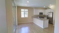 This two-floor space presents a clean, modern aesthetic anchored by an open-concept main living area. The kitchen is outfitted with crisp white cabinetry, granite countertops with subtle veining, and stainless steel appliances including a built-in microwave. A peninsula with a breakfast bar extends into the living space, creating a natural division while maintaining an open, connected feel. Recessed lighting is installed throughout the ceiling, providing consistent illumination across the space.

Light hardwood-style flooring runs throughout the main level, contributing to a bright and airy atmosphere. The walls are finished in a warm neutral tone, complementing the overall palette. A window positioned on the far wall allows natural light to filter in, offering views of surrounding greenery. A sliding glass door to the left provides additional natural light and outdoor access, further enhancing the sense of openness. The layout is practical and well-proportioned, making efficient use of the available space. The second floor likely contains additional living or sleeping quarters, adding to the overall functionality of this multi-level home.