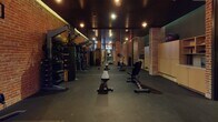 This single-floor fitness space exudes an industrial aesthetic defined by prominent exposed brick walls, dark paneled ceilings, and structural brick columns that anchor the layout. The open floor plan provides a generous training area, with dark rubber flooring extending throughout to support a wide variety of workouts.

The strength training zone is well-appointed with a full cable crossover machine, a rack of medicine balls, a heavy punching bag, multiple adjustable benches, and an extensive dumbbell rack. Full-length mirrors line one wall, amplifying the sense of space and allowing users to monitor their form. Built-in wooden storage cabinetry along one wall offers organized equipment storage.

The cardio section features treadmills and an elliptical machine positioned near a mounted flat-screen television, creating an engaging workout environment. A water cooler and waste receptacle are conveniently placed near the entrance area, alongside private restroom access through dark-framed doors.

Recessed and track lighting casts a warm, focused glow throughout, complementing the brick tones and contributing to a motivating yet intimate atmosphere. The overall design balances raw industrial character with functional, thoroughly equipped training space.