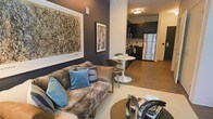 This single-floor space showcases a thoughtfully designed open-concept layout that seamlessly connects the living area, dining nook, and kitchen into one cohesive environment. The living room is anchored by a plush, textured sofa adorned with blue accent pillows, complemented by a round coffee table and a neutral area rug. Bold, large-scale abstract artwork decorates the deep charcoal-toned walls, lending a gallery-like sophistication to the space. A compact dining area features a sleek white pedestal table paired with modern chairs, positioned just steps from the kitchen. The kitchen is outfitted with dark cabinetry, stainless steel appliances including a full-size refrigerator, and ample counter space, all finished with warm wood-look flooring that flows throughout the unit. The consistent use of dark wall tones contrasted with lighter furnishings and metallic accents creates a contemporary, urban aesthetic. Recessed and flush-mount ceiling lighting ensures the space remains well-lit, while the efficient, open arrangement maximizes the sense of spaciousness throughout this well-appointed unit.