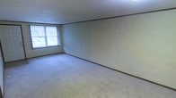 This single-floor space presents a straightforward and functional layout with a neutral, understated aesthetic throughout. The main living area is finished with wall-to-wall carpeting in a light beige tone, providing a soft and comfortable underfoot feel. Smooth, light-colored walls with dark brown baseboard trim run throughout, lending a subtle contrast to the overall palette. The textured popcorn ceiling adds a classic character consistent with the period of construction. Natural light enters the room through a large window dressed with vertical blinds, offering both brightness and privacy control. The space maintains a clean, open feel with minimal architectural ornamentation, making it highly adaptable for a variety of furniture arrangements and uses. The single-floor configuration ensures easy accessibility and a cohesive flow throughout. Overall, the space offers a practical and livable environment with a calm, neutral ambiance well-suited to personalization.