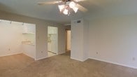 This single-floor residential unit offers a practical and functional layout designed for comfortable everyday living. The main living area is finished with wall-to-wall carpet in a warm neutral tone, creating a cozy and inviting atmosphere. A ceiling fan with a built-in light fixture is centrally mounted overhead, providing both air circulation and ambient lighting. The textured ceiling adds a classic residential touch throughout the space.

An open pass-through connects the living area to the kitchen, which features white cabinetry, white appliances including a refrigerator and range, and light-toned flooring — offering a clean, bright aesthetic. The kitchen is well-organized with ample cabinet storage above and below the counters.

A central hallway extends from the living area, providing access to additional rooms within the unit. HVAC vents are integrated into the walls, ensuring climate control throughout. The walls are painted in a soft, neutral tone that complements the overall light and airy feel of the space. White baseboards frame each room, adding a clean, finished look. The layout is straightforward and efficient, making good use of the available footprint across the single level.