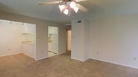 This single-floor residential unit offers a practical and functional layout designed for comfortable everyday living. The main living area is finished with wall-to-wall carpet in a warm neutral tone, creating a cozy and inviting atmosphere. A ceiling fan with a built-in light fixture is centrally mounted overhead, providing both air circulation and ambient lighting. The textured ceiling adds a classic residential touch throughout the space.

An open pass-through connects the living area to the kitchen, which features white cabinetry, white appliances including a refrigerator and range, and light-toned flooring — offering a clean, bright aesthetic. The kitchen is well-organized with ample cabinet storage above and below the counters.

A central hallway extends from the living area, providing access to additional rooms within the unit. HVAC vents are integrated into the walls, ensuring climate control throughout. The walls are painted in a soft, neutral tone that complements the overall light and airy feel of the space. White baseboards frame each room, adding a clean, finished look. The layout is straightforward and efficient, making good use of the available footprint across the single level.