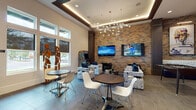 Located at 9010 Running Bull Rd in Orlando, FL, this single-story amenity space blends contemporary design with an energetic, art-inspired aesthetic. The reception area welcomes visitors with large-format tile flooring, a sleek green-accented front desk, and a striking abstract painting that sets the creative tone for the entire space.

The main lounge area is the heart of the facility, anchored by a dramatic full-height exposed brick accent wall adorned with multiple flat-screen televisions. Sculptural decorative lighting cascades from a coffered ceiling with warm LED cove illumination, creating an inviting atmosphere. Patterned upholstered lounge chairs with bold blue accent pillows are arranged around geometric side tables, offering comfortable seating for groups.

Large windows flood the lounge with natural light and frame views of the outdoors, complemented by unique freestanding wooden sculptures that add an artistic focal point. A graffiti-style arcade cabinet adds a playful, urban edge to the gaming corner.

A dedicated corridor features additional curated artwork displayed against the brick wall, with a built-in bench seat and patterned carpet detailing. The space also includes a bowling lane area and additional recreation zones, visible in the dollhouse view. Throughout, the design balances bold artistic expression with functional, social gathering spaces.
