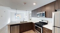This single-floor space showcases a well-appointed kitchen designed with both style and functionality in mind. Rich, dark espresso-toned cabinetry lines the walls, extending to the upper reaches of the space to maximize storage capacity. The cabinetry is complemented by crisp, light quartz countertops that provide ample workspace and a striking visual contrast.

Stainless steel appliances, including a gas range, over-the-range microwave, and dishwasher, lend a sleek, contemporary feel to the kitchen. A double-basin undermount sink is positioned at the peninsula, which also serves as a natural divider between the kitchen and the adjacent living area. Two minimalist pendant lights hang above the peninsula, providing focused task lighting while enhancing the modern aesthetic.

Light-toned hardwood flooring runs throughout, contributing warmth to the space. The open-concept layout allows natural light to flow freely from the living area into the kitchen, with windows visible in the background offering views of the surrounding exterior. The overall ambiance is clean, bright, and modern, making the kitchen both an inviting and highly functional space.