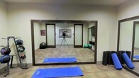 This single-floor fitness studio is designed to support a variety of exercise activities in a functional and straightforward setting. The room features a large, dark-framed wall mirror that spans a significant portion of one wall, allowing users to monitor their form during workouts. The flooring consists of a patterned tile or carpet tile, providing a practical surface for movement. A blue yoga mat is positioned in the foreground, indicating the space is actively used for stretching, yoga, or floor-based exercise. Stability balls are visible in the background near the walls, alongside what appears to be a small shelving unit for equipment storage. Framed artwork on the wall adds a modest decorative touch to the otherwise utilitarian environment. The room is finished with neutral-toned walls and is illuminated by interior lighting, creating a clean and focused atmosphere. Double doors are reflected in the mirror, suggesting convenient access and the potential for the space to accommodate small group classes or individual training sessions. Overall, the studio offers a practical, well-equipped environment for diverse fitness activities.