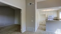 This single-floor residence showcases an immediately striking design element: impressively tall ceilings that lend a sense of openness and airiness throughout the living areas. The interior is finished in a neutral warm-gray palette, complemented by crisp white trim and door casings that create a clean, cohesive aesthetic.

The main living area features soft wall-to-wall carpeting underfoot, contributing to a comfortable and quiet atmosphere. A centrally positioned thermostat and well-placed HVAC vents — both high on the wall and at the baseboard level — point to a thoughtfully designed climate control system that ensures year-round comfort.

An open doorway leads to an adjacent room, also carpeted and finished in the same neutral tones, suitable for use as a bedroom or flexible living space. To the opposite side, a passageway connects to what appears to be a kitchen area, where white upper cabinetry and a countertop overhang are visible, suggesting a functional layout with ample storage. The kitchen benefits from overhead lighting, brightening the space naturally.

Overall, the layout feels connected and practical, with the tall ceilings serving as the standout architectural feature that elevates the ambiance of the entire interior.