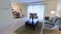 This single-floor residence offers a comfortable and well-appointed living environment with an open layout that seamlessly connects the living and dining areas. The living room is anchored by plush wall-to-wall carpeting and features a large picture window dressed in flowing, floral-patterned curtains that allow abundant natural light to fill the space, creating a bright and airy atmosphere. A dark wood coffee table rests atop a textured green area rug, complementing the neutral tones of the sofa and upholstered armchair arranged nearby. Decorative touches, including a tall vase with sculptural branches, add a refined yet approachable character to the room. The adjoining dining area is furnished with a compact round table and elegantly patterned accent chairs, making it ideal for intimate gatherings. A decorative wall piece adds an artistic focal point to the dining nook. The overall color palette of soft creams and warm beiges lends the space a cohesive, serene ambiance throughout. Recessed lighting provides consistent illumination, while the open flow between spaces enhances the sense of roominess and ease of movement. The thoughtful layout and classic furnishings make this a welcoming and functional single-level home.