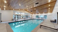 This single-level indoor pool facility combines functional design with a welcoming ambiance. The centerpiece is a spacious rectangular swimming pool with clearly marked lap lanes, its turquoise water reflecting the warm glow of recessed lighting overhead. The vaulted wood-paneled ceiling is a standout architectural feature, adding natural warmth and a resort-like character to the space while amplifying the sense of height and openness throughout the room.

Large black-framed windows and glass-paned doors line the walls, allowing generous natural light to filter in and offering views of the surrounding outdoor environment. This abundance of glazing creates a bright, airy atmosphere that connects the interior to the outdoors without compromising the climate-controlled environment.

The pool deck is finished in light-toned tile and provides ample space for lounging, furnished with chaise lounge chairs and side tables positioned along one side. Additional seating, including chairs and tables, is arranged near the windows on the opposite side, offering a relaxed area for occupants to gather. The overall design strikes a balance between recreational functionality and aesthetic comfort, making it a well-appointed aquatic space.