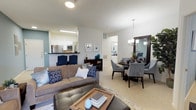 Located at 8101 Fonthill Court in Langhorne, Pennsylvania, this single-floor residence offers a well-designed open-concept layout that seamlessly connects the main living areas. The living room is a welcoming, generously proportioned space featuring plush carpeting, soft neutral tones accented by cool blues, and ample room for comfortable furnishings. A large sofa and coffee table anchor the space, which flows effortlessly into the adjacent dining area. The dining space is illuminated by an elegant chandelier and features a round table with upholstered chairs, making it ideal for both casual meals and entertaining. A large mirror enhances the sense of space and light in this area. The kitchen is visible from the living area and is equipped with stainless steel appliances, including a refrigerator and microwave, along with a raised breakfast bar and crisp white cabinetry. The clean, bright white walls throughout the home lend an airy, open ambiance, while consistent flooring and thoughtful transitions between spaces create a cohesive feel. The overall design balances functionality with comfort, making this a practical and inviting single-level home in the Langhorne, PA community.