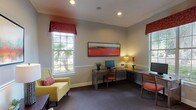 This single-floor space is thoughtfully designed to balance functionality with a welcoming atmosphere. The main area serves as a business center, featuring an L-shaped workstation configuration with multiple computer setups, allowing several users to work simultaneously. Natural light floods the room through large multi-pane windows dressed with rich red floral valances, offering views of mature outdoor greenery. Crown molding, recessed lighting, and a chair-rail detail running along the walls lend the space a refined, polished character. Plush carpeting in a neutral tone grounds the room comfortably underfoot.

A bold, panoramic red abstract artwork anchors the main wall above the workstations, adding a vibrant focal point. A seating area with cheerful yellow armchairs and red accent pillows provides a relaxed spot for conversation or waiting, complemented by a dark wood side table and a warm table lamp. An adjacent lounge connects seamlessly to what appears to be a bar or kitchenette area, visible through an open doorway.

A secondary room off the main space includes an additional workstation, a fold-out sofa in deep navy, and a window overlooking landscaped grounds, making it a versatile flex room suited for private work, rest, or overflow accommodation.