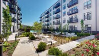 This pool courtyard in Annapolis, Maryland, is a thoughtfully designed outdoor amenity space set within a contemporary residential community. Framed on multiple sides by modern multi-story buildings clad in light gray siding with dark-framed windows and private balconies, the courtyard offers a well-defined, park-like atmosphere that balances architecture with nature.

At the heart of the space, a swimming pool serves as the central focal point, complemented by an array of white outdoor tables and chairs arranged across wide concrete patio areas ideal for lounging and socializing. A striking wood-beamed pergola provides a shaded gathering area and adds warm architectural contrast to the building exteriors. Lush, professionally designed landscaping weaves throughout the courtyard, featuring ornamental grasses, flowering shrubs, boxwoods, and young deciduous trees that lend seasonal color and greenery.

Concrete pathways connect the various zones of the courtyard, leading residents past garden beds to building entry points, including access to a lifeguard room and additional interior amenity spaces visible through large glass doors. Decorative metal privacy screens with intricate branch-like patterns add artistic detail near the pool entry. Pathway lighting fixtures are integrated at ground level, supporting evening use of the space. The open-sky setting allows abundant natural light throughout the day, creating a bright and inviting outdoor retreat.