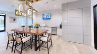 This contemporary single-floor amenity lounge is designed to offer a sophisticated and inviting communal experience. The space is defined by light wood-toned tile flooring that flows seamlessly throughout, creating a warm and cohesive aesthetic.

The lounge area is anchored by a striking black geometric textured feature wall that serves as a dramatic focal point, complemented by a long upholstered banquette adorned with an eclectic mix of colorful accent pillows. Round metal pedestal tables are paired with mustard yellow swivel chairs, while an adjacent seating cluster features tufted blue and gray armchairs arranged atop a vibrant, richly patterned area rug. Illuminated open shelving adds a decorative accent and soft ambient glow to the space.

Floor-to-ceiling black-framed glass doors and windows line the perimeter, flooding the interior with natural light and offering direct views to an outdoor area, enhancing the open and airy atmosphere.

The kitchenette and dining area is equally well-appointed, centering on a live-edge wood communal table surrounded by woven-back chairs beneath a sculptural multi-pendant light fixture. The kitchenette features sleek white cabinetry, a stainless steel sink, dishwasher, and a wall-mounted flat-screen television. Tall paneled storage cabinets contribute a clean, modern finish to the overall design.