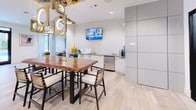 This contemporary single-floor amenity lounge is designed to offer a sophisticated and inviting communal experience. The space is defined by light wood-toned tile flooring that flows seamlessly throughout, creating a warm and cohesive aesthetic.

The lounge area is anchored by a striking black geometric textured feature wall that serves as a dramatic focal point, complemented by a long upholstered banquette adorned with an eclectic mix of colorful accent pillows. Round metal pedestal tables are paired with mustard yellow swivel chairs, while an adjacent seating cluster features tufted blue and gray armchairs arranged atop a vibrant, richly patterned area rug. Illuminated open shelving adds a decorative accent and soft ambient glow to the space.

Floor-to-ceiling black-framed glass doors and windows line the perimeter, flooding the interior with natural light and offering direct views to an outdoor area, enhancing the open and airy atmosphere.

The kitchenette and dining area is equally well-appointed, centering on a live-edge wood communal table surrounded by woven-back chairs beneath a sculptural multi-pendant light fixture. The kitchenette features sleek white cabinetry, a stainless steel sink, dishwasher, and a wall-mounted flat-screen television. Tall paneled storage cabinets contribute a clean, modern finish to the overall design.
