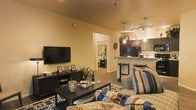 This single-floor residence offers a thoughtfully designed open-concept layout that seamlessly connects the living area and kitchen, creating a cohesive and inviting atmosphere. The living room is comfortably appointed, featuring hardwood-style flooring, a large flat-screen television mounted on a warm-toned accent wall, and a stylish media console. A patterned area rug anchors the seating arrangement, which is dressed in colorful, textured throw pillows that add personality and warmth to the space. Overhead, track lighting provides focused illumination throughout the main living area. The kitchen is positioned just steps away, separated by a breakfast bar with counter-height seating — ideal for casual dining or entertaining. Dark cabinetry contrasts with lighter countertops, while pendant lighting suspended above the bar adds a modern touch. Stainless steel appliances, including a full-sized refrigerator, complete the functional kitchen space. A hallway visible from the living area leads to additional rooms, hinting at a well-organized floor plan. Throughout the space, warm neutral wall colors paired with rich wood tones create an ambiance that feels both modern and homey, making it a comfortable and stylish place to live.