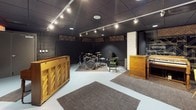 This music room is a purpose-built space designed for rehearsal and recording, offering a well-organized layout across a single floor. The main live room features a dark, acoustically treated environment with a drop ceiling finished in black tile and outfitted with track lighting that provides focused, adjustable illumination throughout the space. Decorative acoustic panels with a woven diamond pattern are mounted along the upper walls, contributing both to sound control and visual character. The room comfortably accommodates a full drum kit, set up on a area rug for additional dampening, alongside vintage wooden cabinet pieces and a keyboard station. A bold black-and-white mural of a guitarist adorns one wall, adding an artistic and inspirational touch to the atmosphere. A separate control or observation room is accessible through a heavy soundproofed door and features a large pass-through window overlooking the live room, along with a built-in wooden credenza with cabinet storage beneath a dark countertop. The floors throughout are finished in light-toned painted concrete, providing a clean contrast to the dark walls and ceiling. The overall ambiance is intimate and functional, with a professional studio feel balanced by eclectic vintage furnishings.