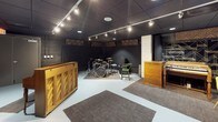This music room is a purpose-built space designed for rehearsal and recording, offering a well-organized layout across a single floor. The main live room features a dark, acoustically treated environment with a drop ceiling finished in black tile and outfitted with track lighting that provides focused, adjustable illumination throughout the space. Decorative acoustic panels with a woven diamond pattern are mounted along the upper walls, contributing both to sound control and visual character. The room comfortably accommodates a full drum kit, set up on a area rug for additional dampening, alongside vintage wooden cabinet pieces and a keyboard station. A bold black-and-white mural of a guitarist adorns one wall, adding an artistic and inspirational touch to the atmosphere. A separate control or observation room is accessible through a heavy soundproofed door and features a large pass-through window overlooking the live room, along with a built-in wooden credenza with cabinet storage beneath a dark countertop. The floors throughout are finished in light-toned painted concrete, providing a clean contrast to the dark walls and ceiling. The overall ambiance is intimate and functional, with a professional studio feel balanced by eclectic vintage furnishings.