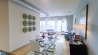 This single-floor space showcases a thoughtfully designed open-plan layout that seamlessly connects the dining and living areas. An exposed metallic ventilation duct runs along the ceiling, lending an industrial character that contrasts beautifully with the otherwise modern, refined aesthetic. The dining area features a sleek glass-top table with chrome X-frame chairs, anchored by a green-striped area rug. A striking cascading bubble chandelier serves as a dramatic focal point overhead. The walls are adorned with a grid of circular green ceramic wall art, adding a bold decorative statement. The living area is furnished with a plush grey sectional and complementary seating, accented with colorful throw pillows. Large windows dressed in sheer white curtains allow generous natural light to flood the space, enhancing the airy, open atmosphere. Light hardwood flooring runs throughout, tying the areas together cohesively. Track lighting and a sleek arc floor lamp provide additional illumination. The overall ambiance is contemporary and eclectic, balancing industrial elements with vibrant décor accents.