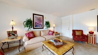 Located on SW 16th St in Ocala, Florida, this single-floor residence offers a comfortable and cohesive layout designed for everyday living. The living room serves as the heart of the home, featuring plush wall-to-wall carpeting, neutral walls, and ample space for relaxed seating arrangements. A large cream sofa, upholstered armchair with ottoman, and a stone-topped coffee table create an inviting atmosphere, complemented by decorative greenery and framed artwork.

Adjacent to the living room, the dining area is anchored by a pendant chandelier that casts warm light over a glass-top dining table surrounded by woven rattan chairs. A bright window allows natural light to filter in, enhancing the room's airy feel.

The kitchen features wood-look flooring, white cabinetry, and essential appliances including a refrigerator and range, with a practical galley-style layout that connects conveniently to the rest of the home. A hallway leads to the laundry area, adding functional convenience.

The bedrooms are carpeted and appointed with classic wooden bed frames featuring ornate headboards, offering a warm and restful retreat. A substantial dark wood armoire in the main living area provides additional storage while serving as a striking design accent. Throughout, the home maintains a cohesive, traditional aesthetic with neutral tones and warm wood furnishings.