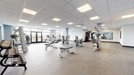 Located at 33 Old Tavern Road in Orange, Connecticut, this fitness center offers a generously sized, single-floor layout designed to accommodate a comprehensive workout experience. The main exercise area is bright and open, featuring a drop ceiling with recessed panel lighting that evenly illuminates the entire space. Durable rubber flooring throughout the workout zone provides both safety and comfort underfoot.

The facility is outfitted with a variety of strength-training machines, adjustable weight benches, and cable equipment, complemented by a dedicated free weights section with a mirrored wall that enhances the sense of openness. Cardio equipment, including treadmills and upright bikes, is positioned near large front-facing windows and glass entry doors, allowing natural light to fill the space and providing views of the surrounding exterior.

Structural columns with a soft blue-gray finish add architectural character while helping define zones within the open floor plan. A separate corridor leads to clean, clearly marked restroom facilities for both men and women, keeping support amenities conveniently accessible yet distinct from the main workout area. The overall design is clean, modern, and functional, creating a welcoming environment well-suited for fitness and wellness use.