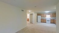This single-floor residence offers a clean, functional layout designed for comfortable everyday living. The main living area is spacious and open, finished with neutral wall tones and soft, light-colored carpeting that lends a warm and inviting atmosphere. Ceiling-mounted lighting and HVAC vents are thoughtfully integrated throughout, ensuring consistent comfort across the space.

Adjacent to the living area, the kitchen is efficiently designed with medium-toned wood cabinetry providing generous storage and counter space. A full-size refrigerator anchors the kitchen, which is well-lit and easily accessible from the main living zone, making it ideal for both daily use and casual entertaining.

A built-in closet with double doors is conveniently situated between the living area and the rest of the unit, offering practical storage solutions. A connecting hallway leads to additional rooms beyond, suggesting a well-organized floor plan that maintains a clear separation between living and private spaces.

Overall, the unit presents a straightforward, move-in-ready layout with a neutral palette throughout, making it easy to personalize. The open flow between the living area and kitchen, combined with ample built-in storage, reflects a practical and efficient use of the available space.