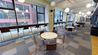 This contemporary single-floor commercial space is thoughtfully designed to accommodate a variety of collaborative and social activities. The layout is divided into distinct functional zones that flow seamlessly into one another, creating an open yet organized environment. Large, floor-to-ceiling windows line the perimeter, flooding the interior with natural light and offering expansive views of the surrounding urban streetscape. Accent walls in bold navy blue and soft sage green add visual interest and help define different areas within the space. Pendant globe light fixtures hang throughout, providing warm, ambient illumination that complements the natural light. The main gathering area features a mix of round café-style tables paired with contemporary wood-and-chrome chairs, alongside bar-height seating along the window ledges — ideal for informal meetings or individual work. Lounge areas are furnished with sleek black leather sofas, low coffee tables, and side tables, creating comfortable spots for relaxed conversation or presentations, supported by wall-mounted displays and a projection screen. The flooring transitions between warm hardwood and patterned carpet tiles with orange accent details, subtly delineating each zone. A quieter, more intimate lounge nook offers a cozy retreat within the larger open floor plan. Overall, the space strikes a balance between energetic openness and purposeful comfort.