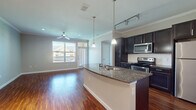 This single-floor residence offers a well-designed open-concept layout that seamlessly connects the kitchen and living areas. Rich hardwood-style flooring extends throughout the main living spaces, complemented by crown molding and neutral-toned walls that create a clean, cohesive aesthetic. Ceiling fans are installed in both the living area and bedroom, contributing to year-round comfort.

The kitchen is a standout feature, centered around a curved granite-topped island with an undermount sink and pendant lighting overhead. Dark espresso cabinetry provides ample storage, while a full suite of stainless steel appliances—including a range, microwave, refrigerator, and dishwasher—adds a modern touch. Track lighting above the kitchen enhances both function and style.

The living area benefits from natural light through large windows with views to the outdoors, and direct access to an exterior door. The bedroom features plush carpeting, an arched window, and a ceiling fan. The bathroom is well-appointed with a granite vanity, dark cabinetry, tile flooring, a tub-shower combination, and convenient access to a walk-in closet.
