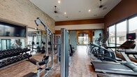This fitness center in Spring, TX, is a thoughtfully designed, single-level facility that offers a comprehensive workout environment across a generously sized open floor plan. The space features a striking blend of exposed brick walls, warm earth-toned accent walls in deep ochre and brown, and durable tile flooring, creating an industrial yet inviting aesthetic.

The cardio zone is lined with Matrix treadmills, ellipticals, and stair climbers, positioned along floor-to-ceiling windows that offer views of an outdoor pool area and surrounding community, providing natural light and a motivating backdrop. Mounted flat-screen televisions are positioned throughout for entertainment during workouts.

The strength training area houses a robust selection of Matrix cable and multi-functional machines, a free-weight section with a full dumbbell rack, and a dedicated functional training zone equipped with kettlebells in various weights and stationary cycling bikes. Large mirrors enhance the sense of space in the weight area.

Soaring ceilings with ceiling fans and recessed lighting ensure proper ventilation and visibility throughout. A distinctive sliding barn-style door serves as a stylish interior partition, adding a modern-rustic design touch. The layout allows ample room for movement between equipment zones, supporting a comfortable and efficient workout experience.
