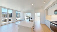 This contemporary 2-bedroom, 2-bathroom residence offers a thoughtfully designed single-floor layout with a clean, modern aesthetic throughout. Light hardwood flooring flows seamlessly across all rooms, complementing the bright white walls and recessed lighting that create an airy, open atmosphere.

The combined kitchen and living area serves as the heart of the home, flooded with natural light from expansive floor-to-ceiling windows and sliding glass doors that open to an outdoor balcony with urban views. The kitchen is outfitted with flat-front white cabinetry extending to the ceiling for ample storage, stainless steel appliances including a refrigerator and microwave, and a sleek center island with a white quartz countertop and matte black faucet.

The primary bedroom is generously proportioned and features large windows framing urban skyline views, while the second bedroom offers similar bright, clean finishes. Both bathrooms are finished in a modern style, with one featuring white subway tile, a glass-enclosed walk-in shower, and matte black fixtures. A dedicated walk-in closet with built-in wire shelving systems provides organized storage. The in-unit laundry area houses a stacked Whirlpool washer and dryer, accessible through a louvered door for added convenience.