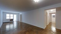 This single-floor space offers a clean, functional layout with timeless design elements throughout. The main living area is immediately striking for its expansive, open feel, defined by rich parquet wood flooring that adds warmth and classic character to the room. Bright white walls provide a neutral, versatile backdrop, while flush ceiling lighting ensures the space is well-illuminated throughout the day and evening. Natural light filters in through a glazed door or window at the far end of the main area, contributing to an airy and welcoming ambiance. The open floor plan allows for flexible furniture arrangements and a natural flow between spaces. Doorways lead to adjoining rooms, suggesting a well-connected layout that maximizes usability across the floor. A hallway visible to one side hints at additional rooms, providing a sense of depth and practicality to the overall floorplan. The clean lines, neutral palette, and durable flooring combine to create a space that is both functional and easy to personalize, making it well-suited for a variety of uses and living arrangements.