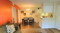 This single-floor residential space offers a thoughtfully designed open-concept layout that seamlessly connects the living, dining, and kitchen areas. The dining area serves as a central focal point, highlighted by an elegant chandelier overhead and hardwood-style flooring that extends throughout the main living spaces. A bold orange accent wall adds a vibrant pop of color, complementing the neutral tones found throughout the rest of the space and creating a visually dynamic atmosphere. Framed botanical artwork adorns the walls, contributing to the warm and inviting character of the home. The dining area comfortably accommodates a full dining set, making it well-suited for everyday meals and entertaining. Adjacent to the dining space, the kitchen features white cabinetry, a breakfast bar with counter seating, and under-cabinet lighting that brightens the workspace. The open pass-through design between the kitchen and dining area enhances the sense of connectivity and spaciousness. The living area, partially visible from the dining space, features plush carpeting and ample room for comfortable seating. Overall, the space balances functionality with a cozy, lived-in aesthetic, making it an approachable and practical single-floor home.