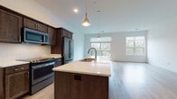 This single-floor residence at 11130 N Buntrock Ave in Mequon, Wisconsin offers a thoughtfully designed open-concept layout finished with contemporary materials throughout. The kitchen serves as the heart of the space, featuring rich dark-stained cabinetry, stainless steel appliances including a gas range, built-in microwave, and refrigerator, along with a herringbone tile backsplash and light quartz countertops. A central island with an undermount sink and elegant pendant lighting above anchors the kitchen and provides a natural transition into the expansive living area. The living space is bathed in natural light from large windows and a sliding glass door that opens onto a private balcony with views of surrounding greenery. Light wood-tone flooring flows seamlessly throughout the main living areas, lending the space a clean, modern aesthetic. The bedroom offers plush carpeting, a window with natural views, and direct access to a generously sized walk-in closet with built-in shelving. The bathroom is finished with tile flooring, subway-tiled walls in the shower enclosure fitted with a sliding glass door, dark cabinetry, and chrome fixtures, creating a refined and functional space. Crisp white walls and an open floor plan contribute to the bright, airy ambiance throughout.
