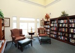 This inviting library and reading room is designed to offer a comfortable and refined retreat for reading and quiet reflection. The space features soaring ceilings with elegant crown molding, lending an open, airy quality to the room. Warm yellow walls create a welcoming atmosphere, complemented by soft grey carpeting throughout.

Multiple dark mahogany bookshelves line the walls, filled with a diverse and extensive collection of books, ranging from novels to reference volumes. A traditional writing desk with brass lamp serves as a functional centerpiece, accompanied by additional smaller bookshelves nearby. Framed botanical prints adorn the walls, adding a classic, scholarly aesthetic to the space.

The seating area is anchored by two plush wingback chairs upholstered in rich earth tones, each paired with a teal leather ottoman for added comfort. A pedestal side table with a brass lamp sits between the chairs. Large multi-pane windows bathe the seating area in generous natural light, offering views of the outdoors.

A glass-paneled door leads to adjacent corridor spaces, suggesting this library is part of a larger facility. Handrails visible through the doorway indicate accessibility-conscious design throughout.