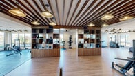 Located on Briargate Trail in Colorado Springs, CO, this modern fitness center offers a thoughtfully designed, single-floor layout that accommodates a wide range of exercise disciplines. The space is defined by its striking architectural details, including a distinctive slatted wood beam ceiling adorned with stylish dome pendant lights, which create a warm yet energetic ambiance throughout.

The main fitness area features light hardwood-style flooring and is divided into functional zones. A dedicated cardio section houses a row of treadmills and elliptical machines positioned alongside floor-to-ceiling windows, flooding the space with abundant natural light and offering views of the surrounding Colorado Springs landscape. Adjacent to this is a strength training zone with rubber tile flooring, equipped with cable machines, weight benches, and free weights.

A central walnut wood open-shelving unit serves as both a stylish design element and a practical divider, storing yoga mats, medicine balls, kettlebells, and other fitness accessories. A separate studio area provides an open, flexible floor plan ideal for yoga, stretching, or group classes. Smart mirror displays and wall-mounted fitness screens are integrated throughout. Large glass-framed doors and interior windows maintain visual connectivity across all zones, contributing to an open, cohesive, and professional fitness environment.
